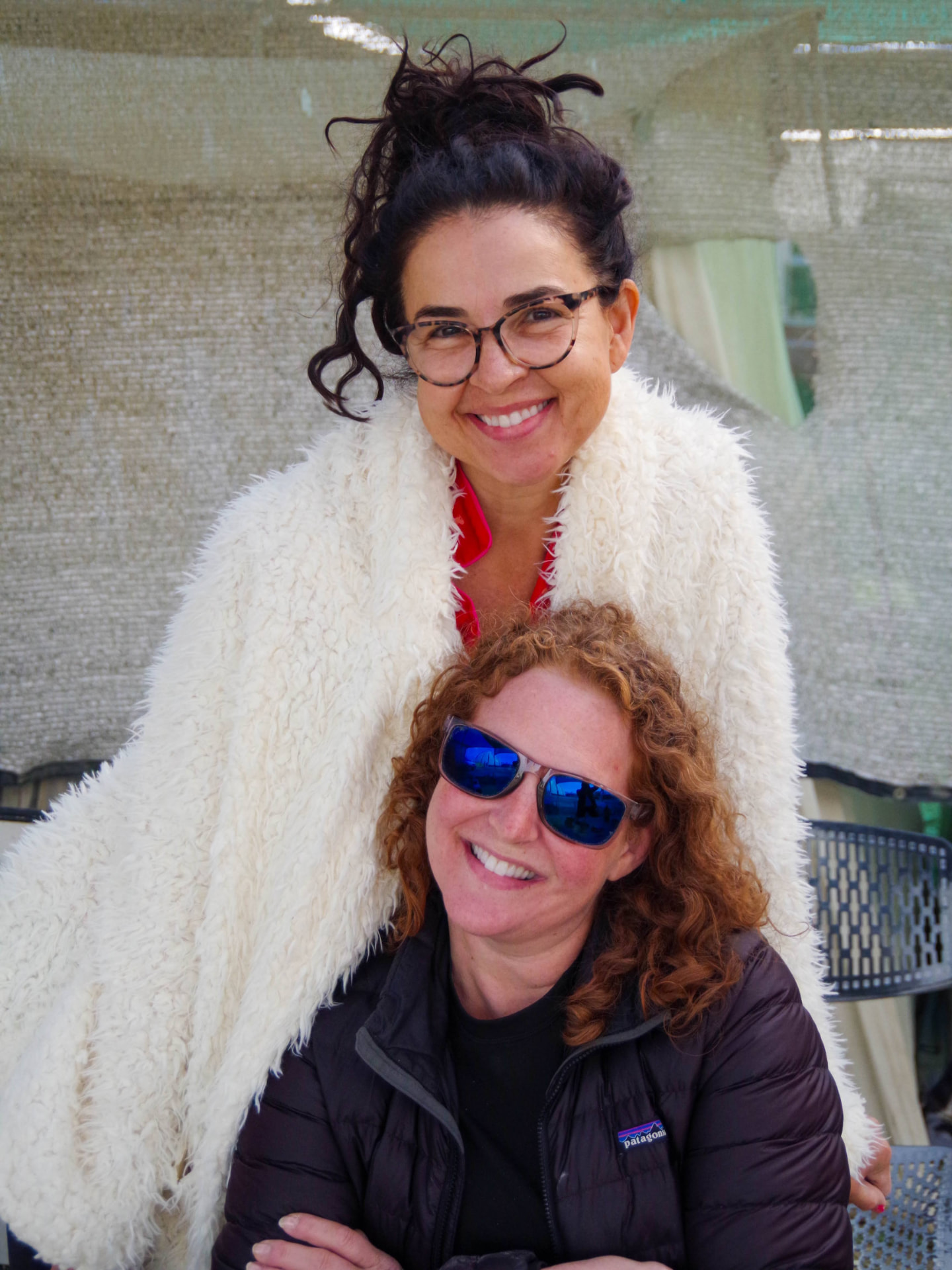 Two campers smiling together under the shade structure, one wrapped in a white fur coat