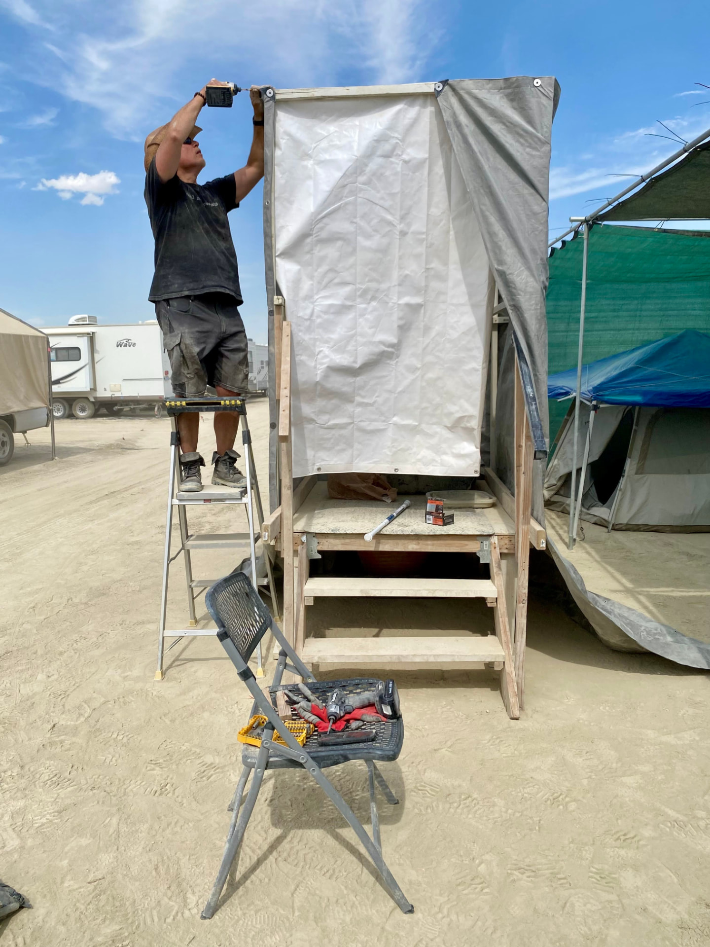 A camper on a ladder building the roof of a wooden shower stall