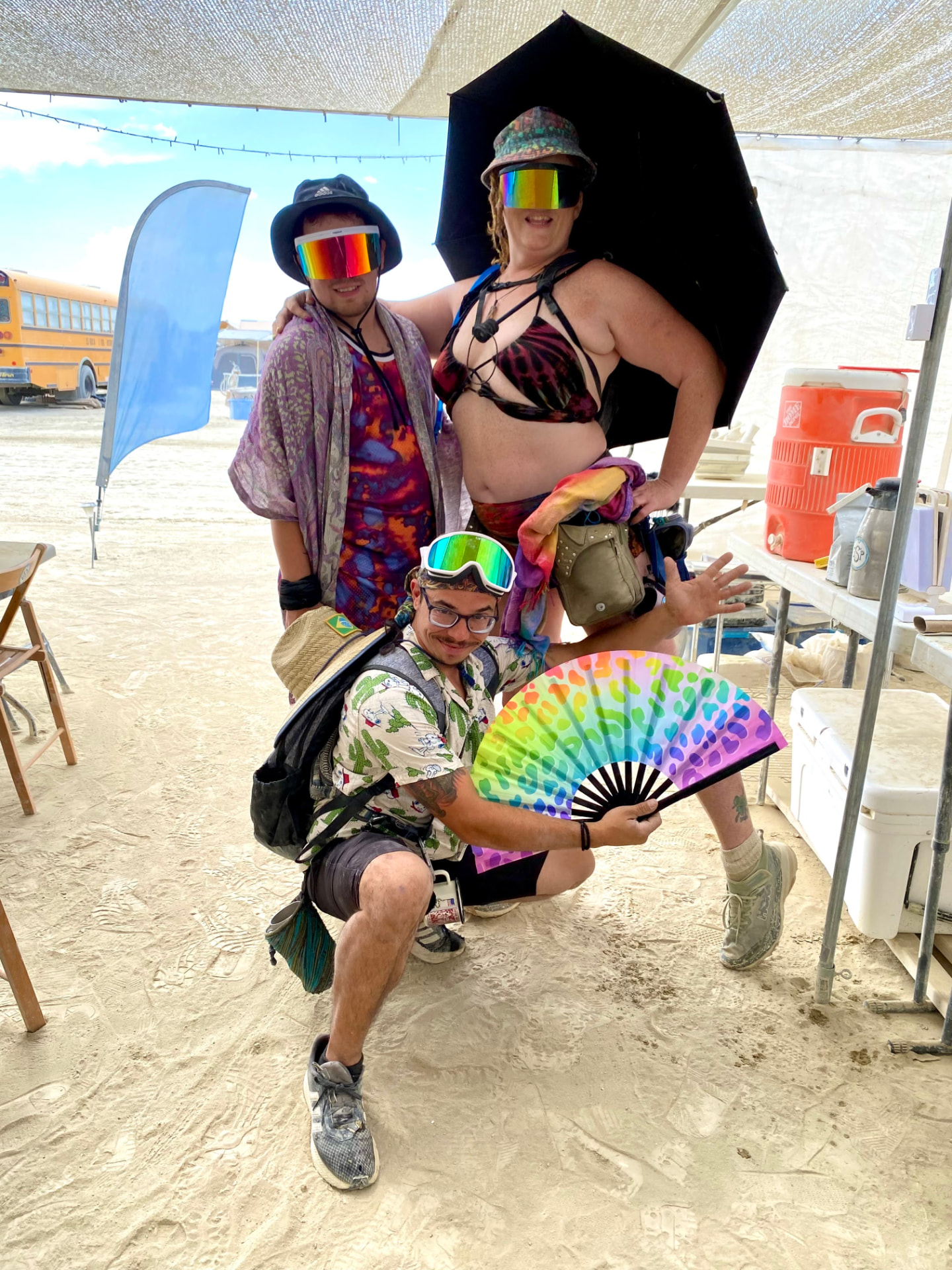 Three campers in colorful outfits and rainbow goggles posing with a large rainbow fan